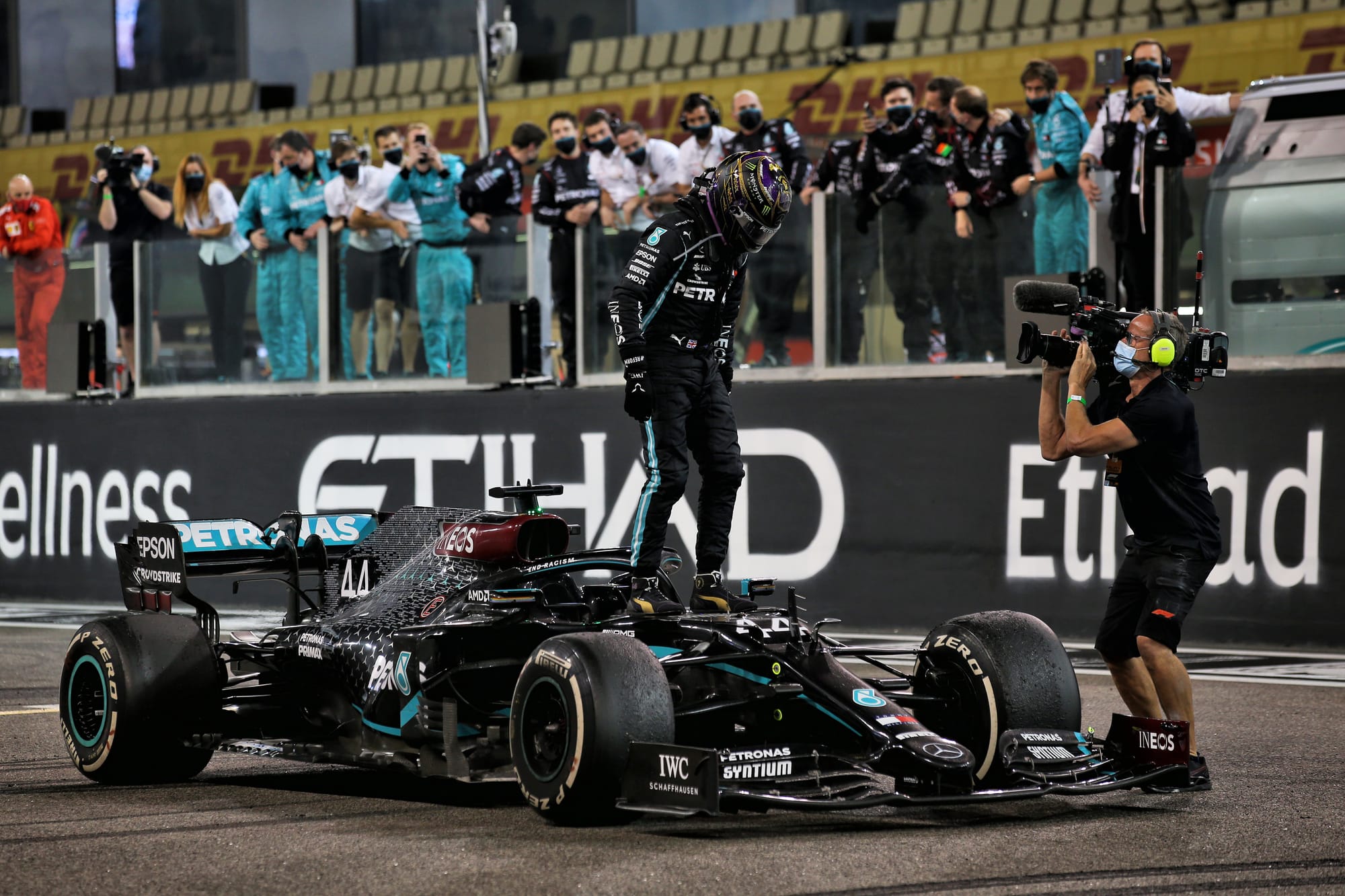 Lewis Hamilton stands on top of the black Mercedes W11 at the final race of 2020 as a cameraman films him on the right-hand side and team members look on in the background